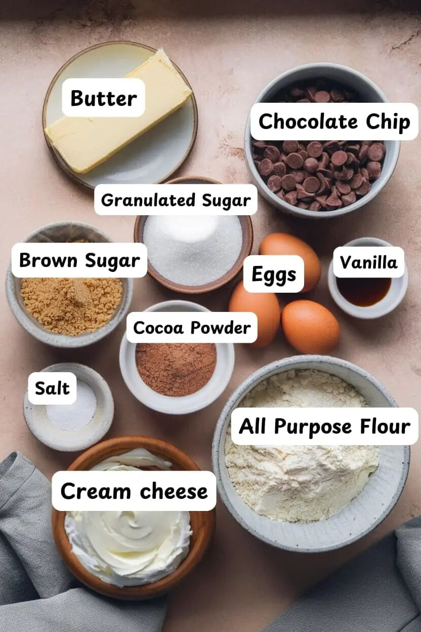 The ingredients of cheesecake brownies on a light brown table including butter, chocolate chips, granulated sugar, eggs, vanilla, brown sugar, cocoa powder, salt, all-purpose flour, and cream cheese.