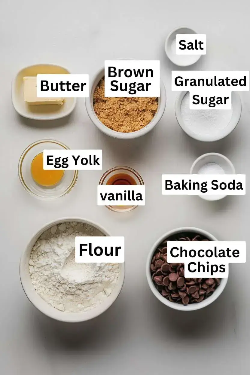 Ingredients on a white surface including salt, brown sugar, butter, granulated sugar, egg yolk, vanilla, baking soda, flour, and chocolate chips.