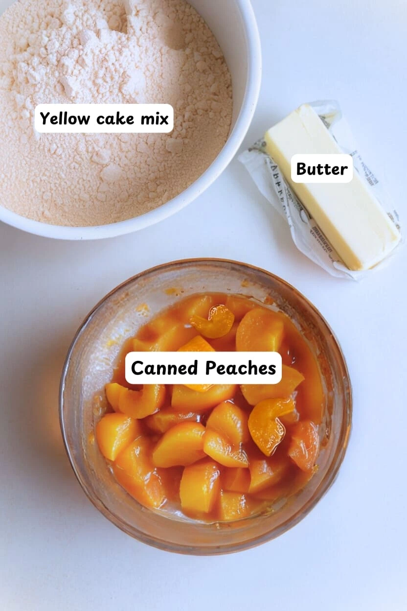 Ingredients for peach cobbler recipe on a white table including canned peaches on a bowl, butter, and yellow cake mix.