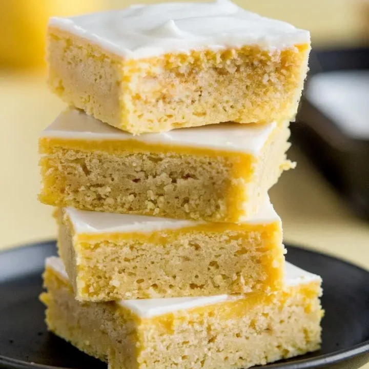 stack of lemon brownies on a black plate atop a light-yellow surface.