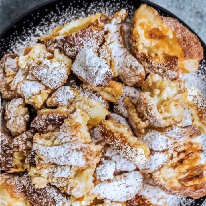 Scrambled pancakes on a black plate on a gray surface with powdered sugar on top.