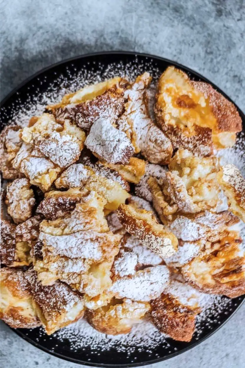 Scrambled pancakes on a black plate on a gray surface with powdered sugar on top.