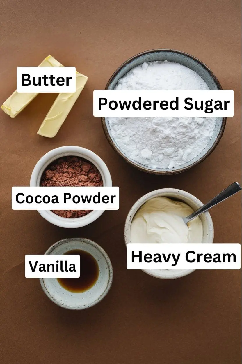 Ingredients of the chocolate frosting on a brown surface including butter, powdered sugar, cocoa powder, heavy cream, and vanilla.