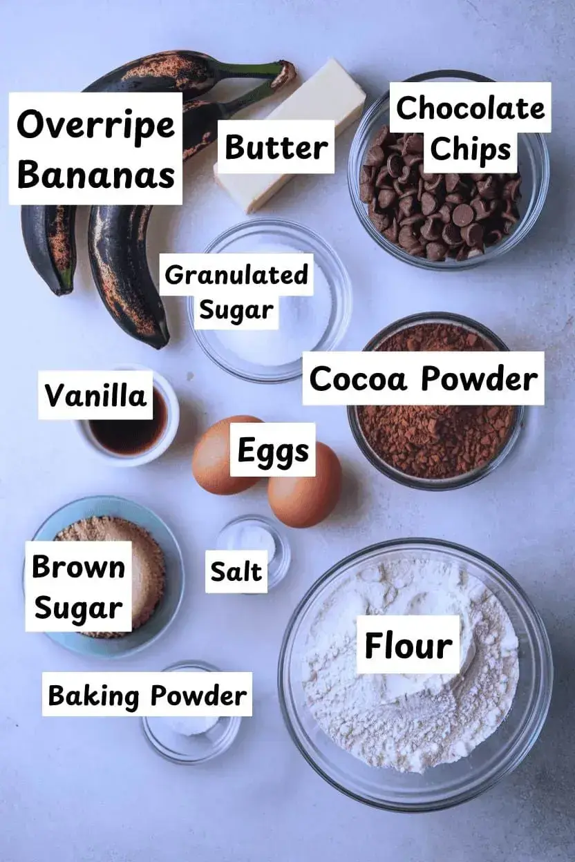 Ingredients for banana brownies on a light gray surface including two overripe bananas, one stick of butter, chocolate chips, granulated sugar, brown sugar, two eggs, vanilla extract, cocoa powder, flour, baking powder, and salt.