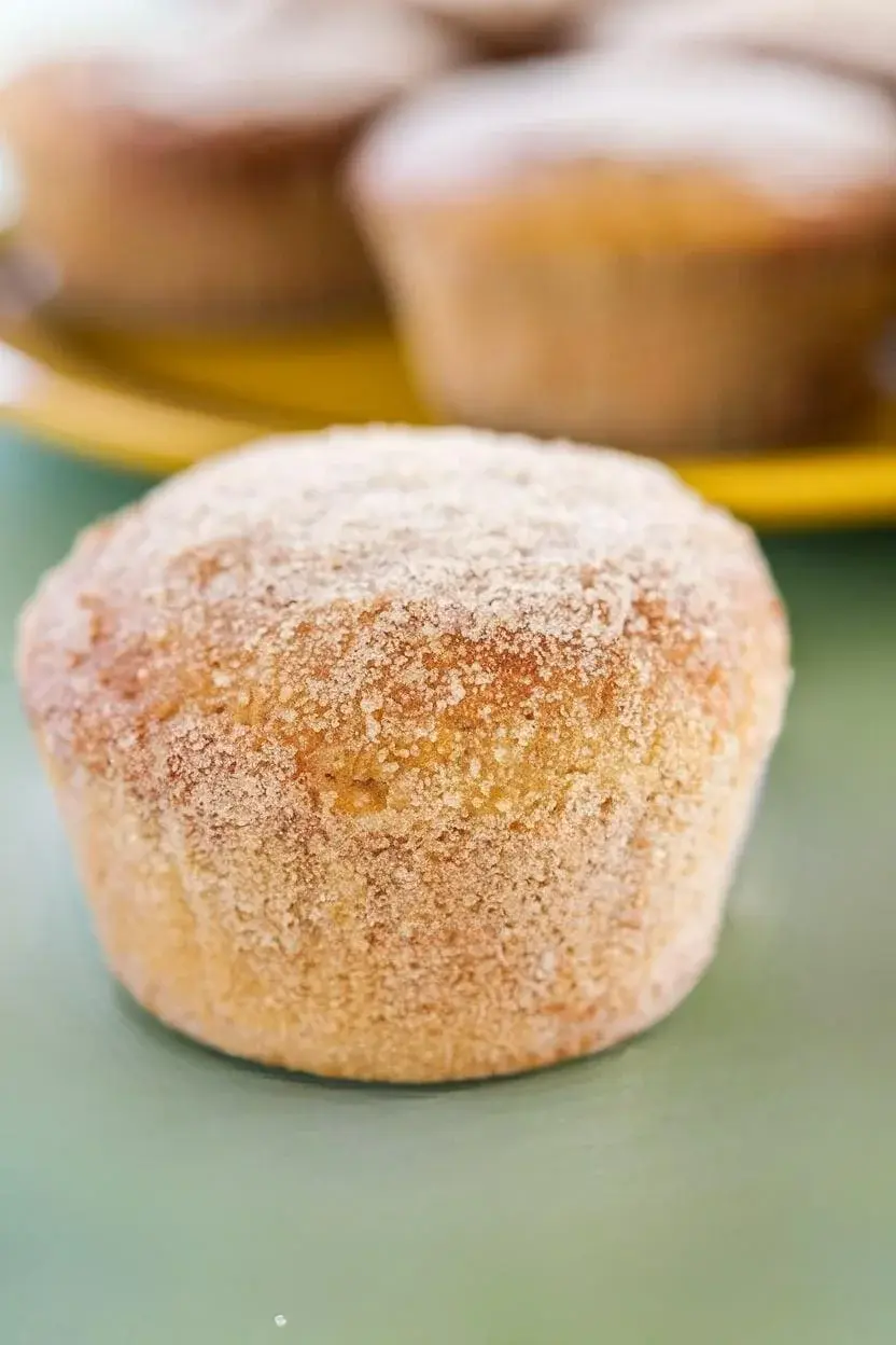 One vegetarian cinnamon sugar donut muffin on a light green surface, with a yellow plate filled with muffins behind it.