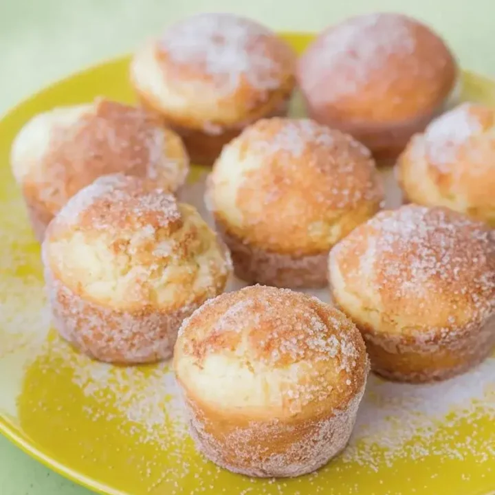 Vegetarian cinnamon sugar donut muffins, coated in cinnamon sugar, are arranged on a yellow plate atop a light green surface.