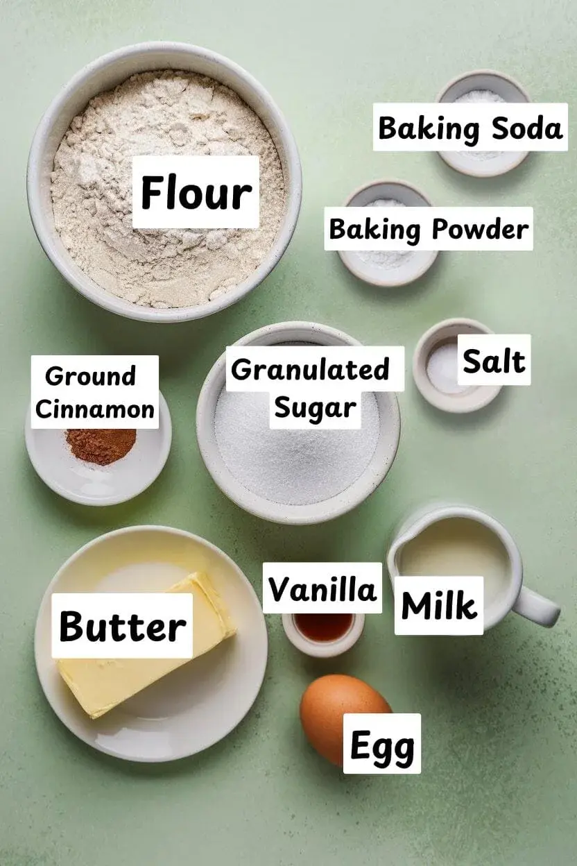 Ingredients on a light green surface including flour, baking powder and baking soda, salt, granulated sugar, ground cinnamon, vanilla extract, butter, milk, and one egg.