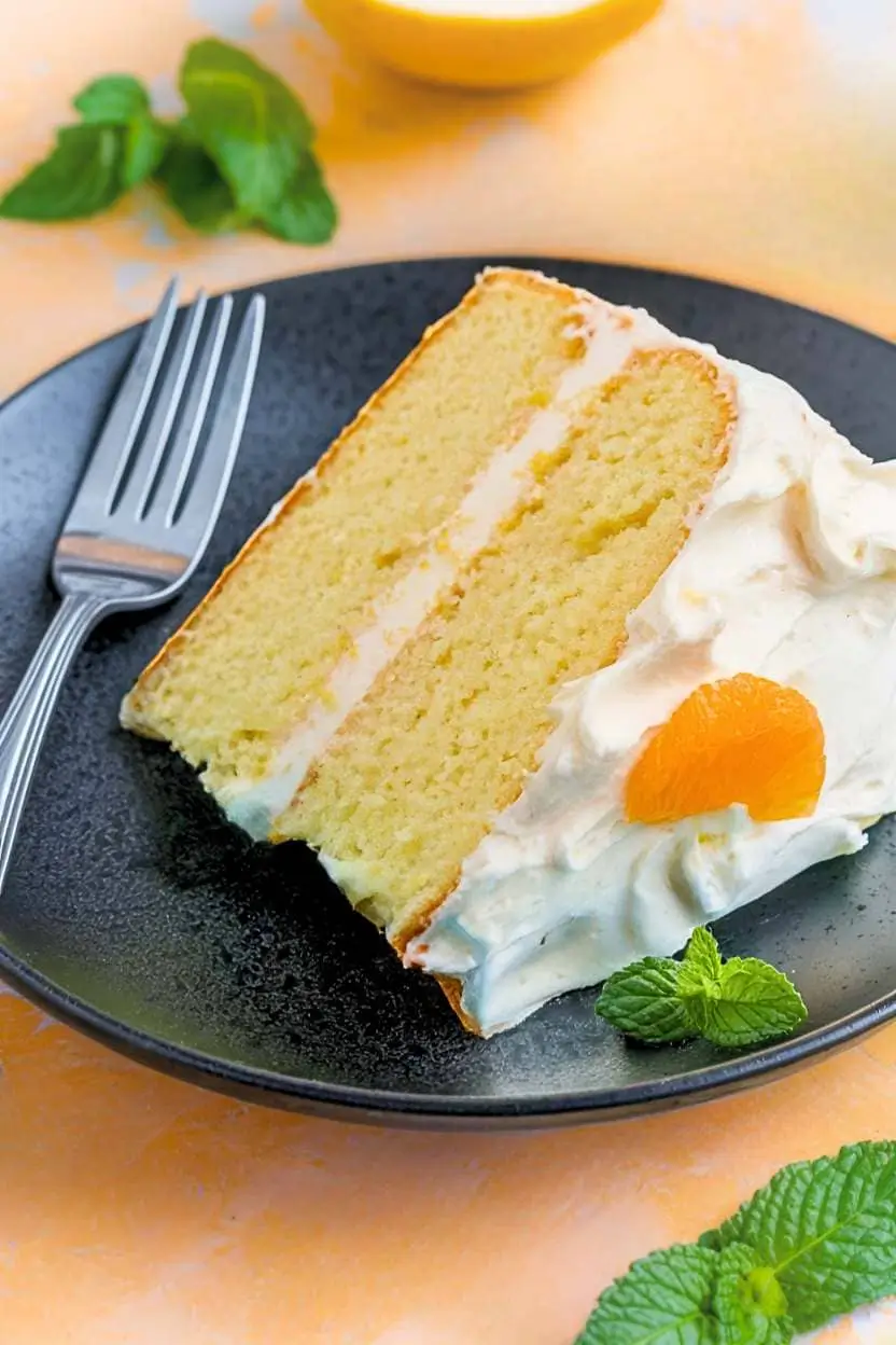 A slice of mandarin orange cake with a fork beside it rests on a black plate against a light orange surface, with some mint around.